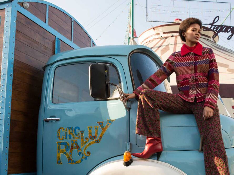 Mujer con estilo afro sentada sobre el capot de un camión antiguo del Circo Raluy Legacy posando para una producción de moda. La mujer lleva un traje estampado colorido y botas rojas. El camión es de color celeste y dice "circ raluy" en la puerta. Eventos. Experiencias.