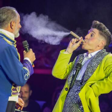 Pietro soplando cigarro de polvo en la cara a Bigotis, el presentador del circo Raluy Legacy. Escena divertida y singular. En el fondo público riendo.