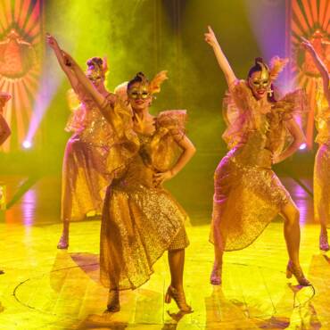 Grupo de mujeres bailando en la pista del Circo Raluy Legacy con luces amarillas y humo. Las mujeres llevan mascaras.