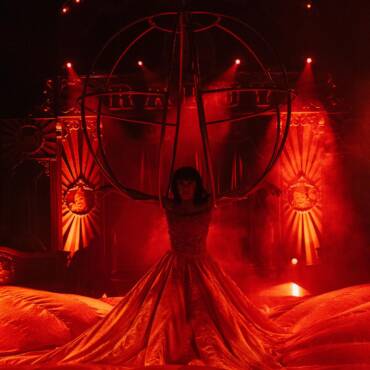 Louisa Raluy con un imponente vestido rojo en un espectáculo del Circo Raluy Legacy, demostrando equilibrio y fuerza al colgarse de un candelabro gigante de metal.