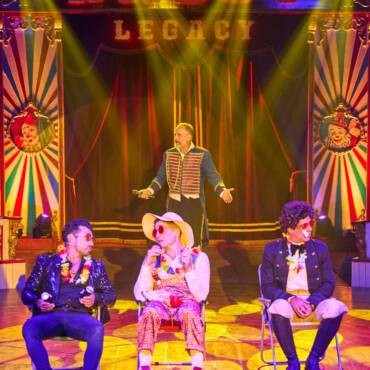 Rodrigo, Pietro y Dimitri sentados en sillas de playa, en el espectáculo del Circo Raluy Legacy. Detrás de ellos está Bigotis, el presentador del circo.