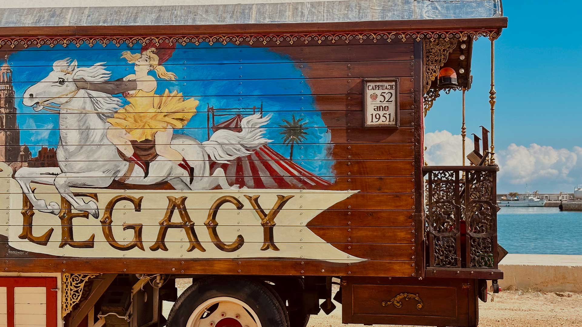 Antiguo carruaje de la caravana del circo Raluy Legacy pintado con mujer con vestido amarillo sobre caballo blanco saltando por encima del logo del circo.