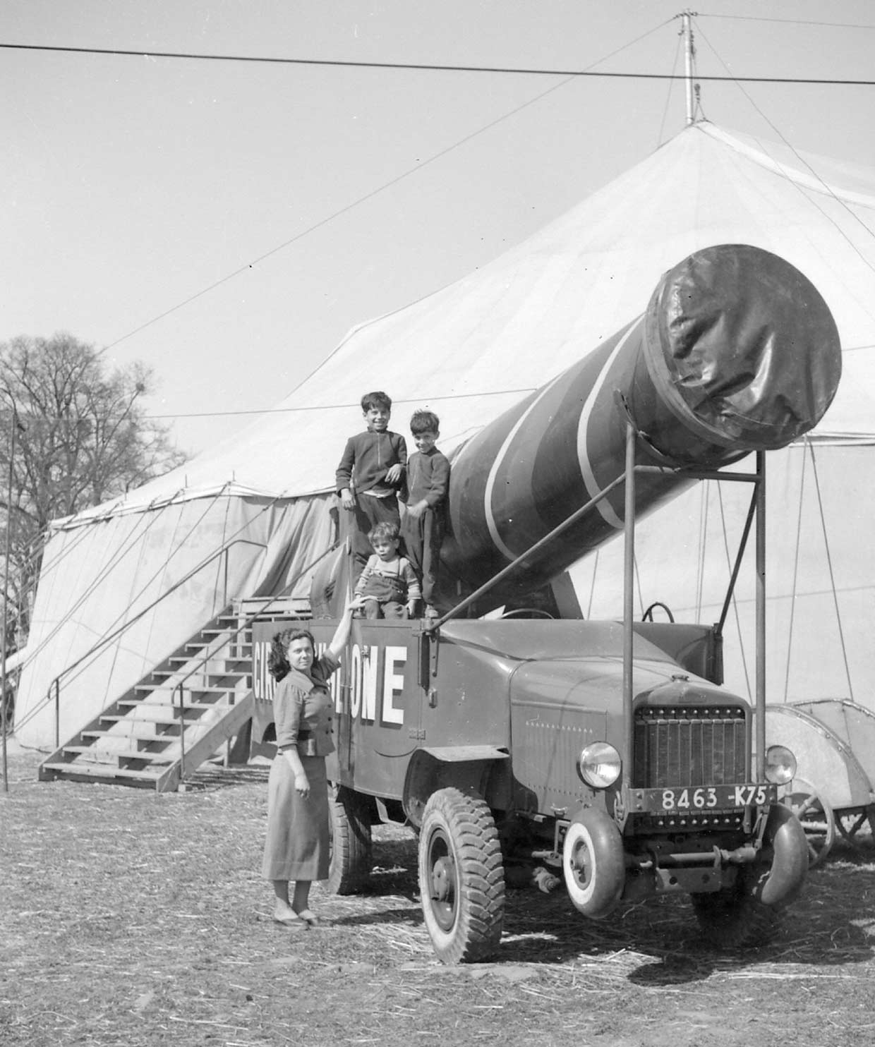 Foto antigua de Marina Tomás junto a un camión que transporta un cañón gigante de circo, delante de una gran carpa blanca. Tres niños está subidos al camión.