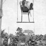 Foto antigua en blanco y negro de Luis Raluy Iglesias haciendo equilibrio sentado en una silla sobre un trapecio. Debajo dos niños mirándolo y de fondo una casa rodeada de vegetación.