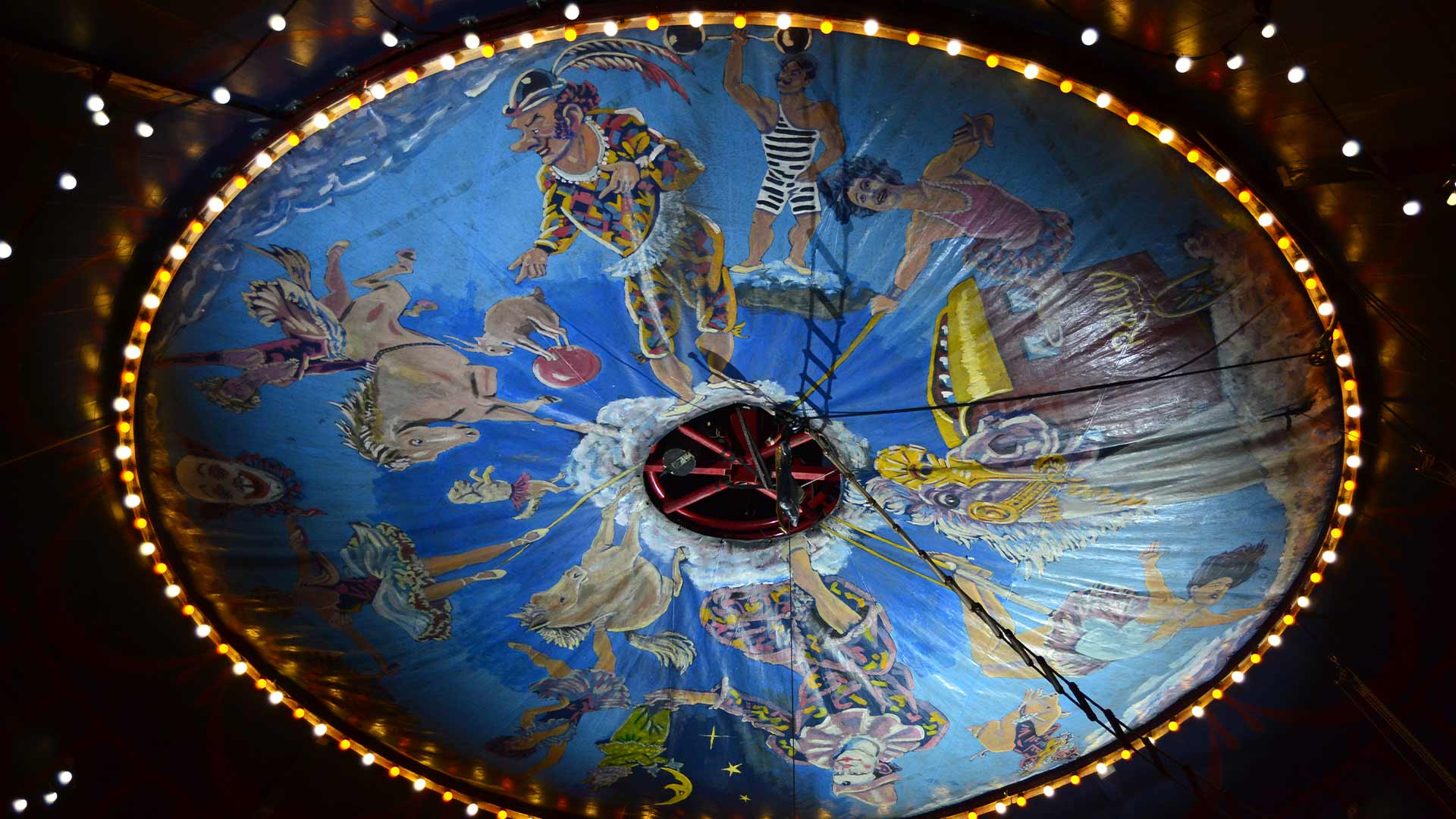 Pintura circular con personajes circenses rodeada de pequeñas bombetas de luz en el techo de la carpa del Circo Raluy Legacy.